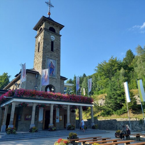 Kamienny kościół z wieżą zegarową, ozdobiony kwiatami i flagami, z ławkami na zewnątrz w górskim otoczeniu