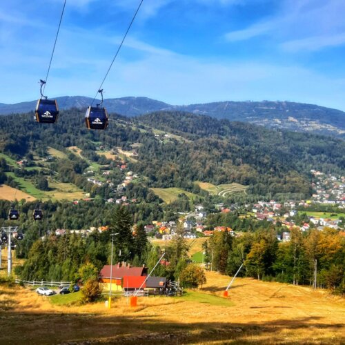 Widok na górskie szczyty z kolejką gondolową nad łąkami i lasem, domki i malownicza wiejska zabudowa.