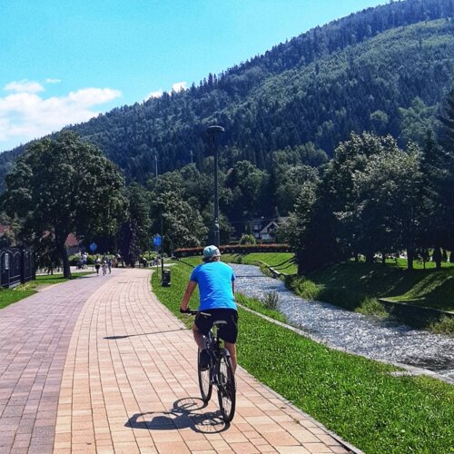 Ścieżka rowerowa nad górskim potokiem z zielonymi drzewami i pagórkami w słoneczny dzień.