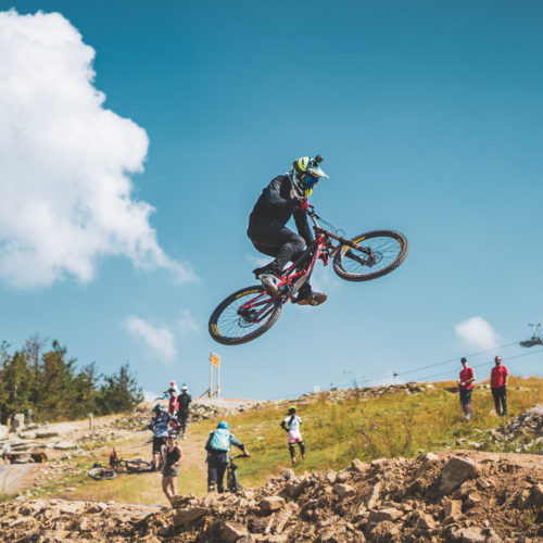 Rowerzysta w kasku wykonuje skok na rowerze górskim na szlaku z widokiem na wyciąg krzesełkowy i las
