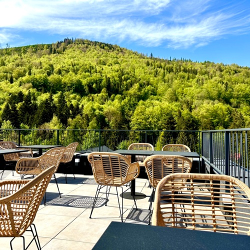 Taras hotelowy z nowoczesnymi rattanowymi krzesłami i stołami z widokiem na zielone, zalesione wzgórze pod błękitnym niebem.