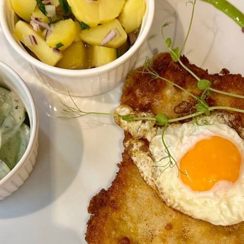 Chrupiący kotlet schabowy z sadzonym jajkiem, podany z sałatką ogórkową i ziemniakami z cebulką i koperkiem.