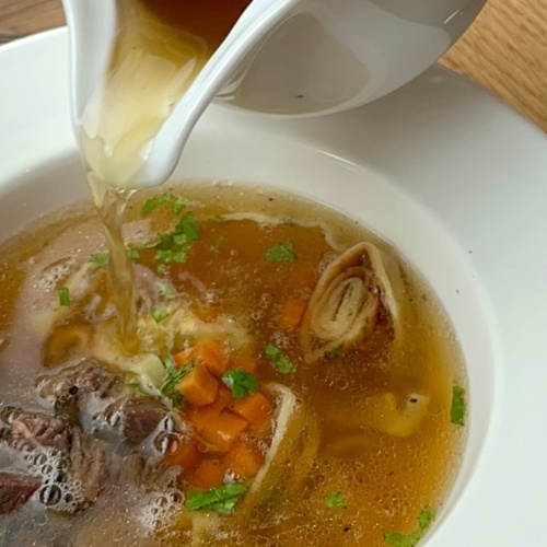 Gorący bulion wlewany do miski z warzywami, mięsem i świeżą natką pietruszki