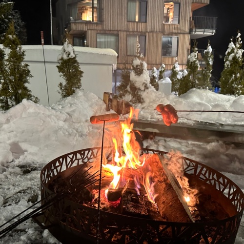 Ognisko w metalowym palenisku na śniegu z pieczonymi kiełbaskami wieczorem przy drewnianym hotelu