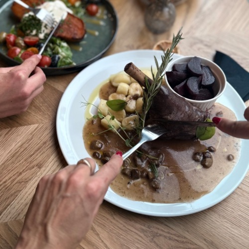 Jídelníček hotelové restaurace s dušeným jehněčím, gnocchi a řepou, host krájí maso vidličkou a nožem.