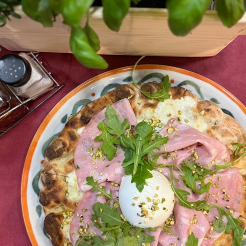 Pizza z dřevěné pece s šunkou, rukolou a kuličkou burraty na keramickém talíři vedle čerstvé bazalky.