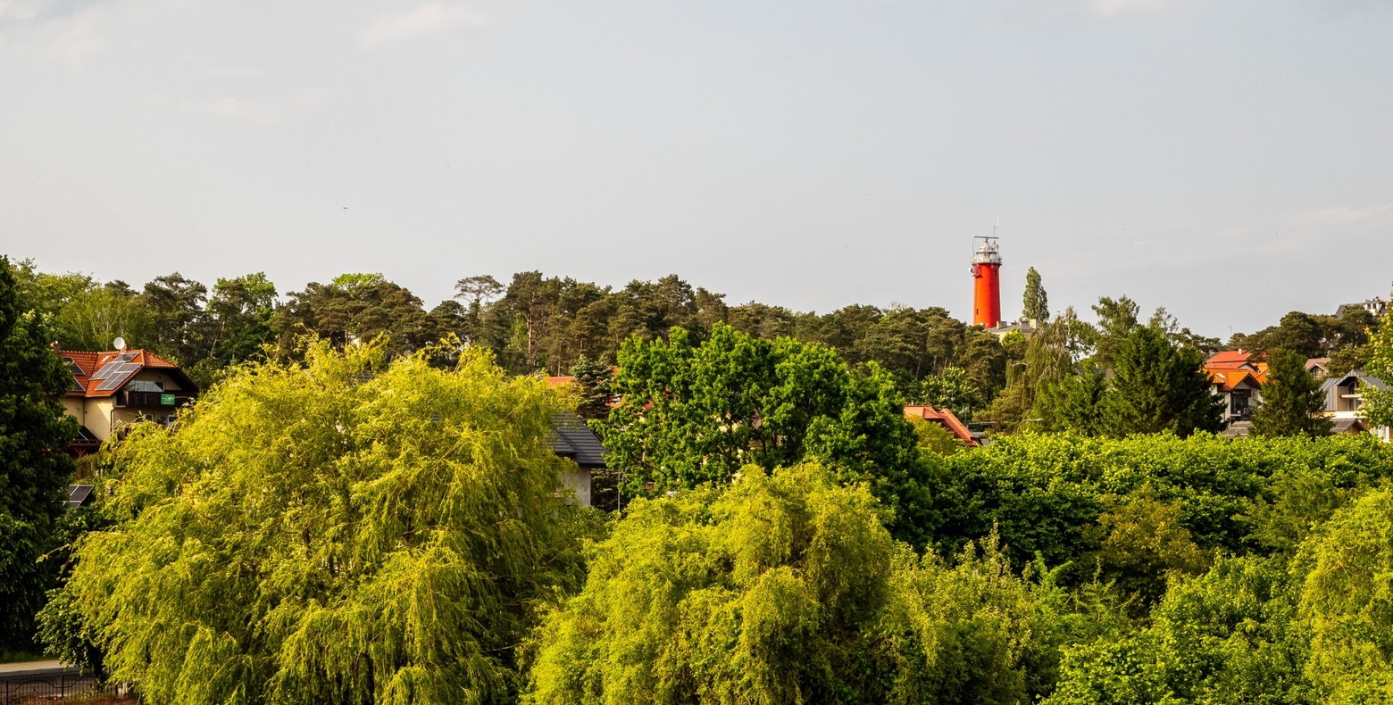 Czerwona latarnia morska ponad gęstą zieloną linią drzew, z rozproszonymi domami i dachami pośród bujnej letniej zieleni