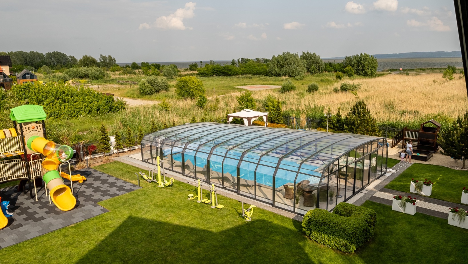 Glass-enclosed hotel pool with loungers, adjacent bright playground slides, outdoor fitness equipment, landscaped lawn and coastal sea view.