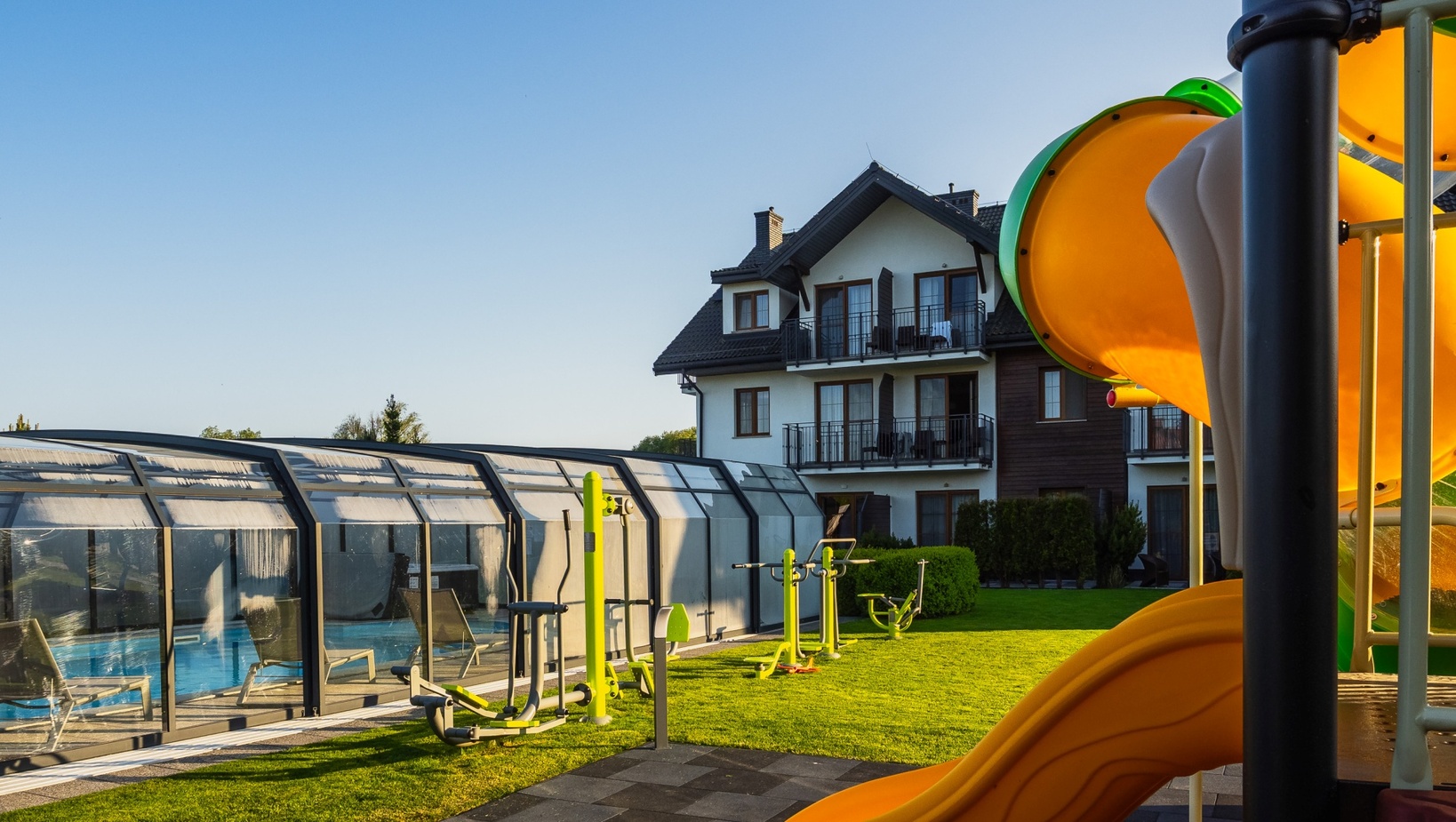 Sunlit family hotel grounds with enclosed glass pool, outdoor fitness machines on the lawn, and bright orange children's slide by guest balconies.