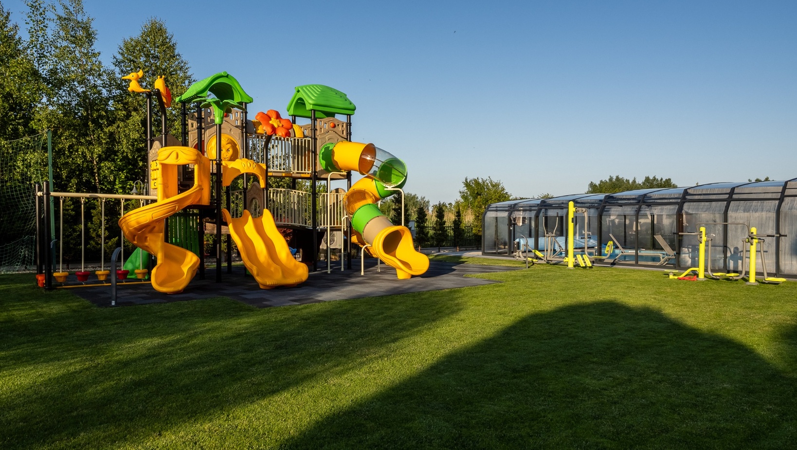 Plac zabaw dla rodzin z żółtymi spiralnymi i rurowymi zjeżdżalniami, zielone daszki nad trawnikiem obok zadaszonego basenu i siłowni