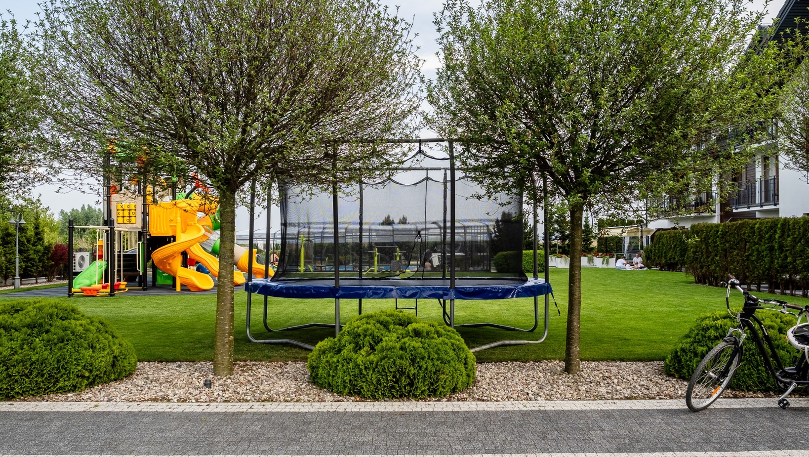 Zielony trawnik hotelowy z ogrodzonym trampolinem, kolorowym placem zabaw, rowerami przy ścieżce i gośćmi piknikującymi przy tarasie.