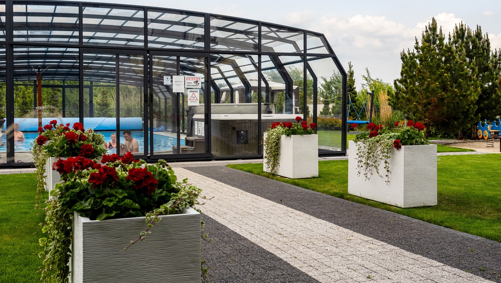 Glass-enclosed heated pool with hot tub and guests swimming, framed by paved walkway, manicured lawn, red flower planters and playground
