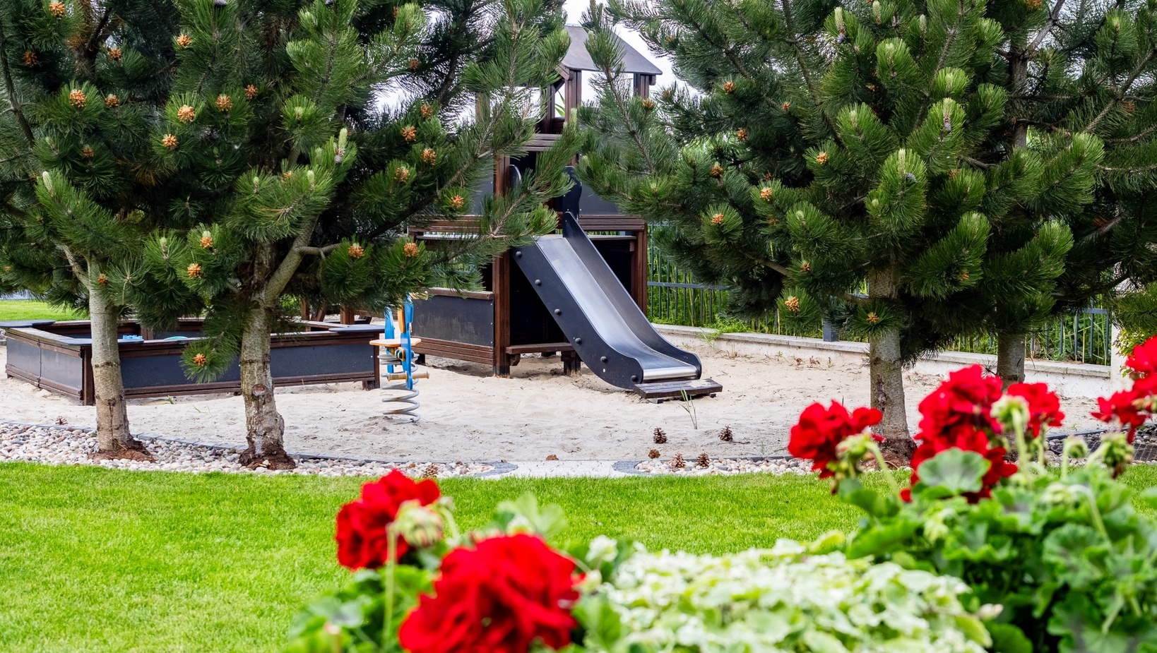 Plac zabaw przy hotelu z czarną zjeżdżalnią i piaskiem, osłonięty sosnami, obok zielonego trawnika i czerwonych pelargonii.