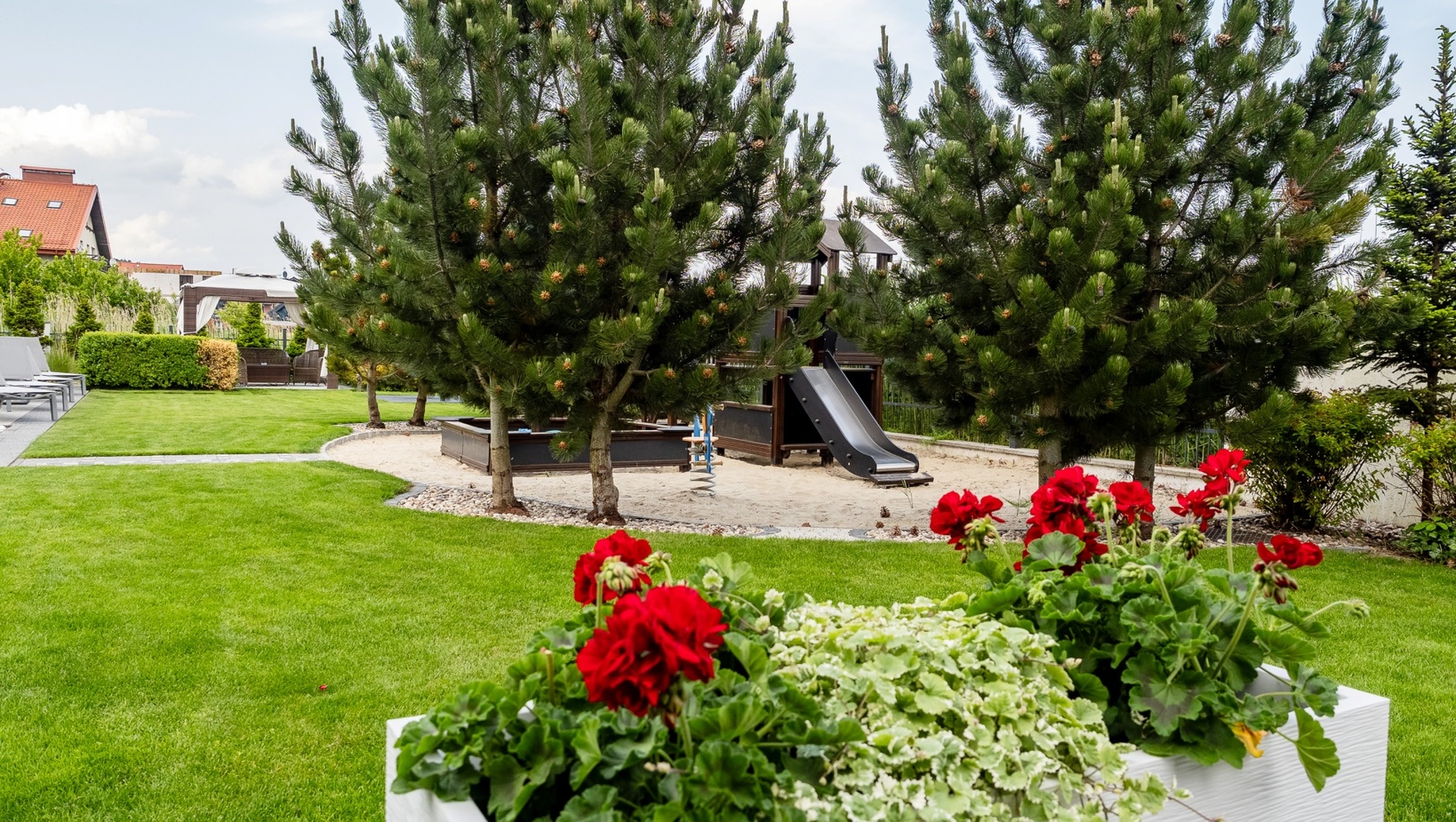 Ogród hotelowy z bujnym trawnikiem, czerwonymi pelargoniami, piaskownicą ze zjeżdżalnią pod sosnami oraz pawilonem i leżakami