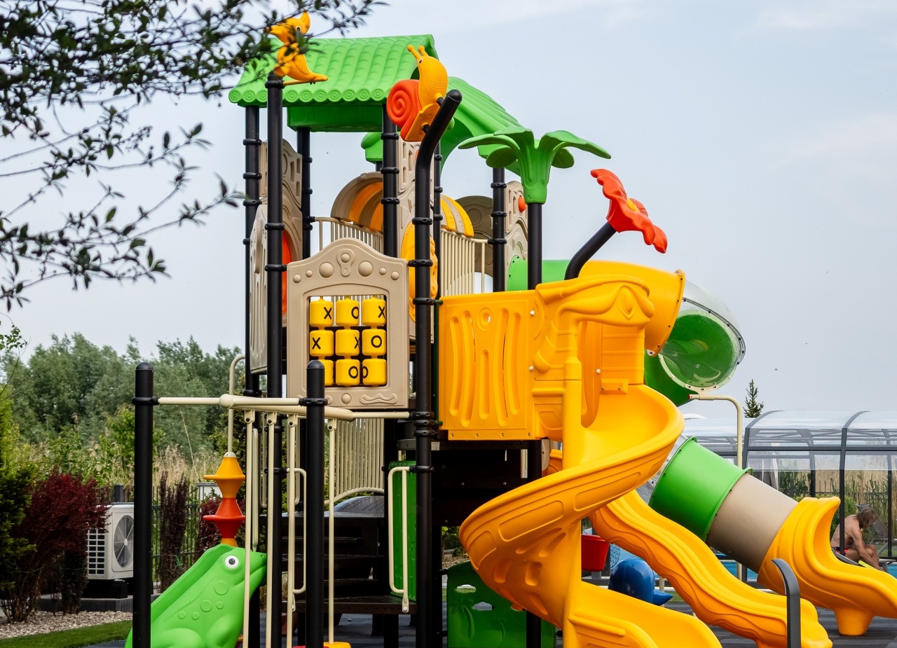 Kolorowy zewnętrzny plac zabaw dla dzieci z zielonym daszkiem, żółtą spiralną i tubową zjeżdżalnią, panelem kółko‑krzyżyk i sztuczną trawą.