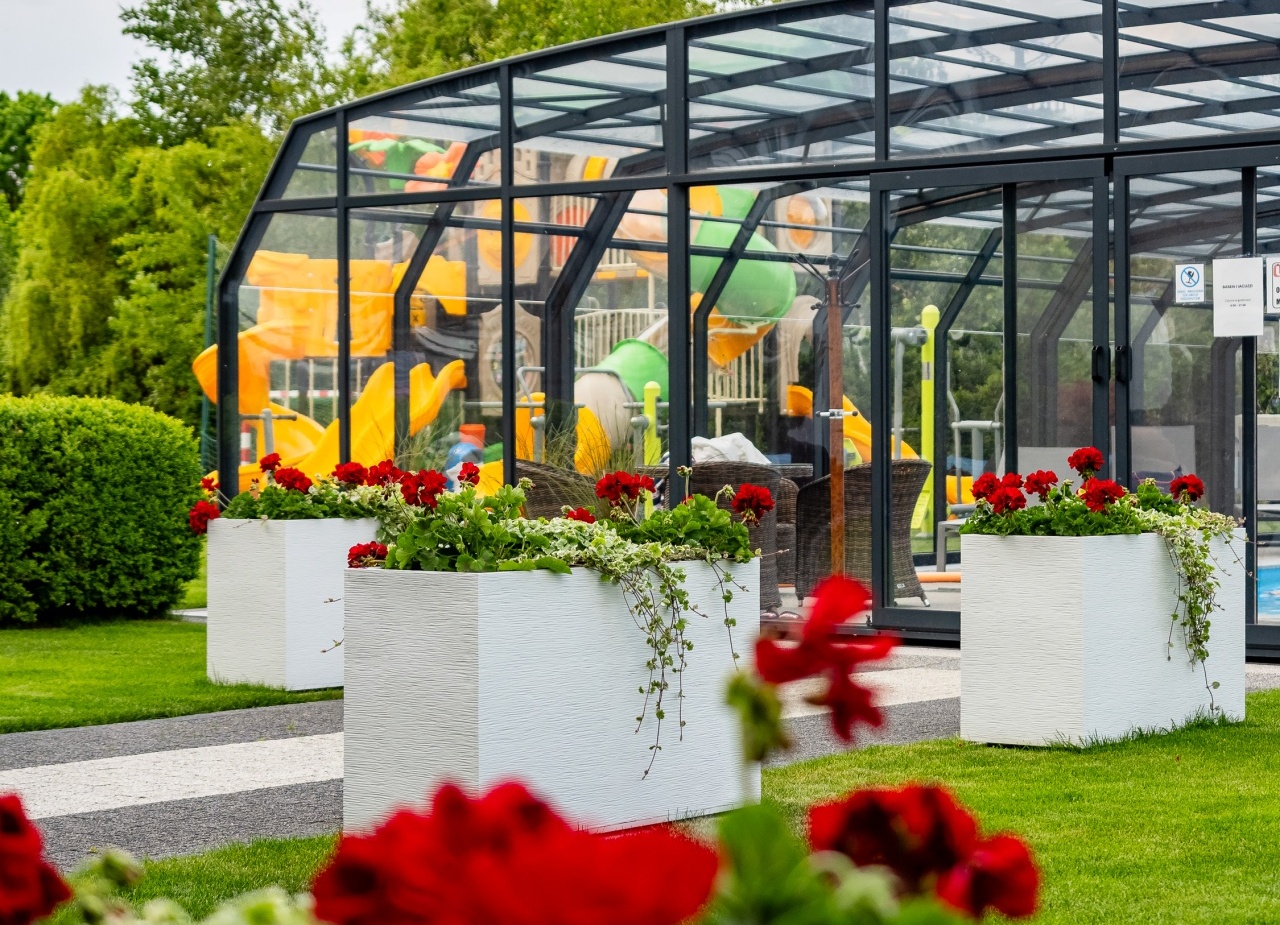 Szklana hala basenowa z kolorowymi zjeżdżalniami, zadbanym trawnikiem i białymi donicami z czerwonymi pelargoniami — atrakcja dla rodzin