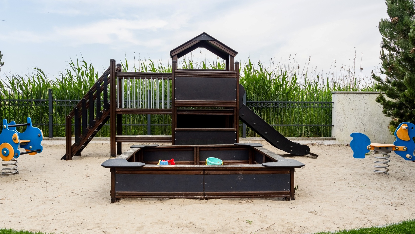 Piaszczysty plac zabaw dla dzieci z drewnianym domkiem i zjeżdżalnią, piaskownicą z zabawkami i dwoma niebieskimi bujakami.