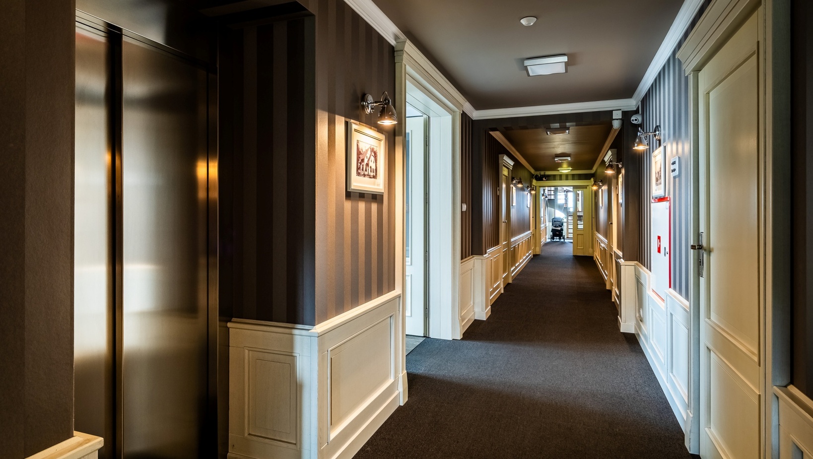Elegancki hotelowy korytarz z wykładziną, białą boazerią, pasiastą tapetą, kinkietami, oprawionymi zdjęciami, windą i wózkiem na końcu