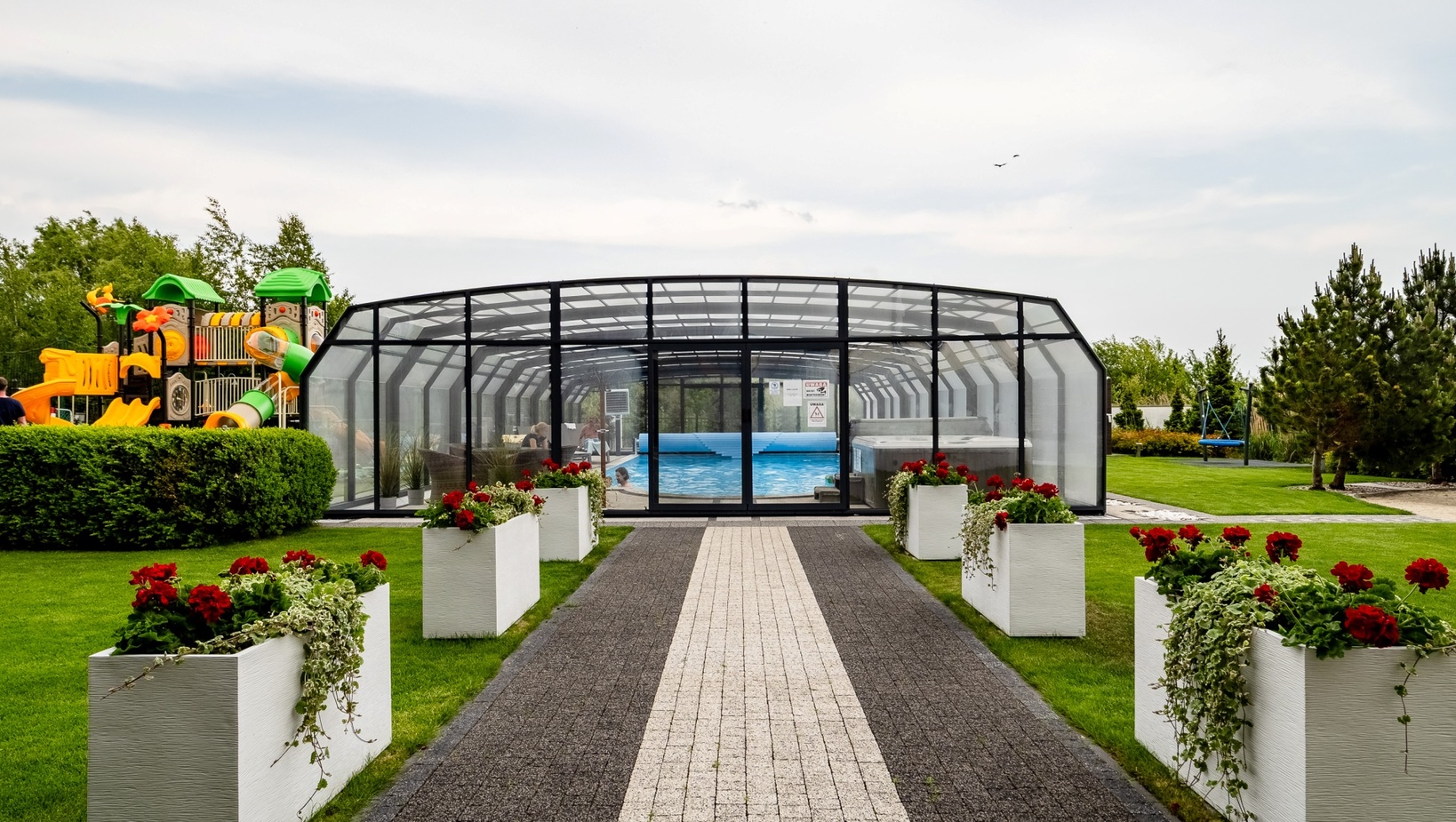 Covered glass-enclosed pool with lounge seating, central paved path lined by white planters with red flowers and a nearby children's playground
