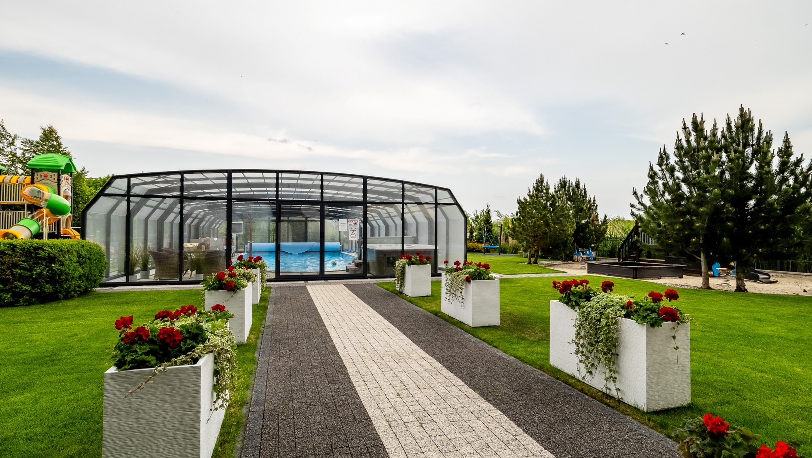 Paved walkway with white planters and red flowers leading to a glass-enclosed pool; green lawns, pines and children's playground.