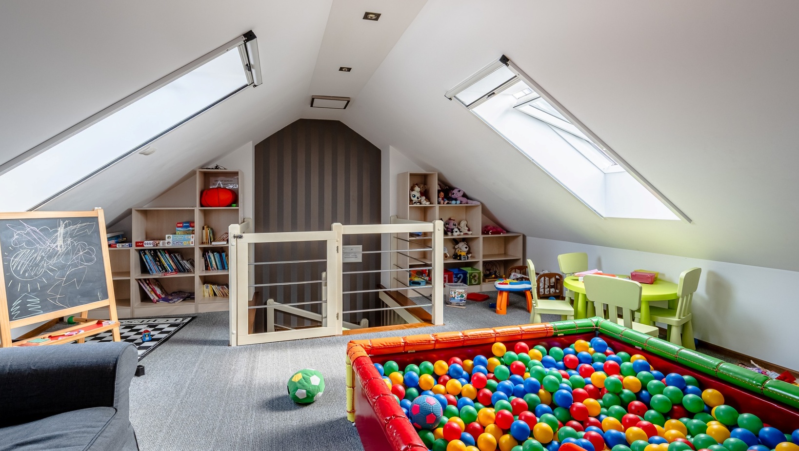 Jasny pokój zabaw na poddaszu z basenem kulkowym, oknami dachowymi, stolikiem i półkami na zabawki