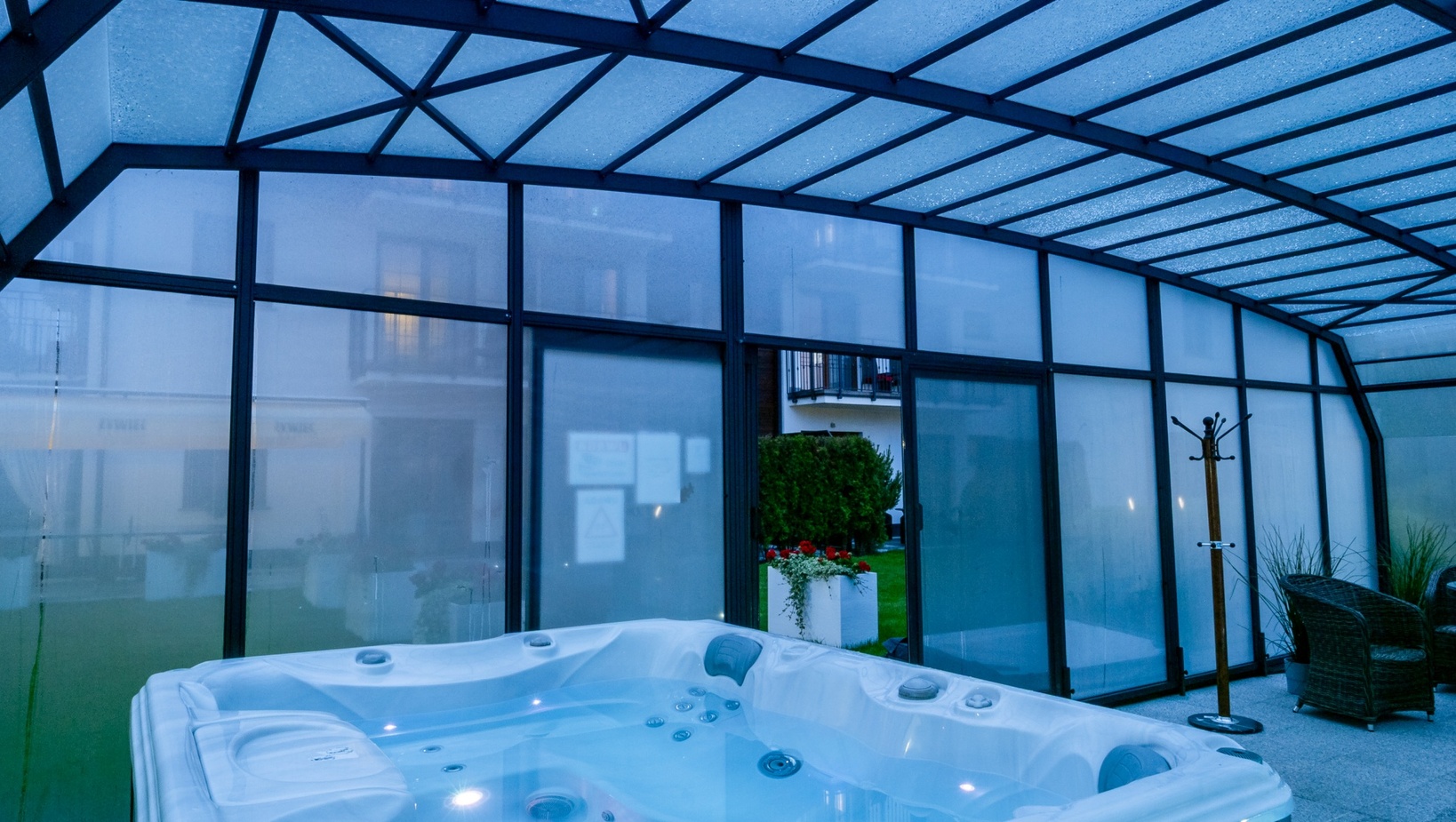 Enclosed glass atrium with illuminated hot tub, wicker seating, coat stand and garden view