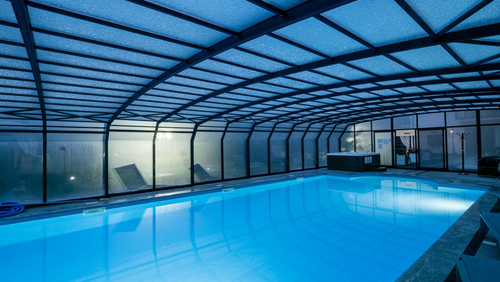 Heated indoor pool with blue ambient lighting, arched glass enclosure, lounge chairs and a hot tub for relaxed spa-style evenings