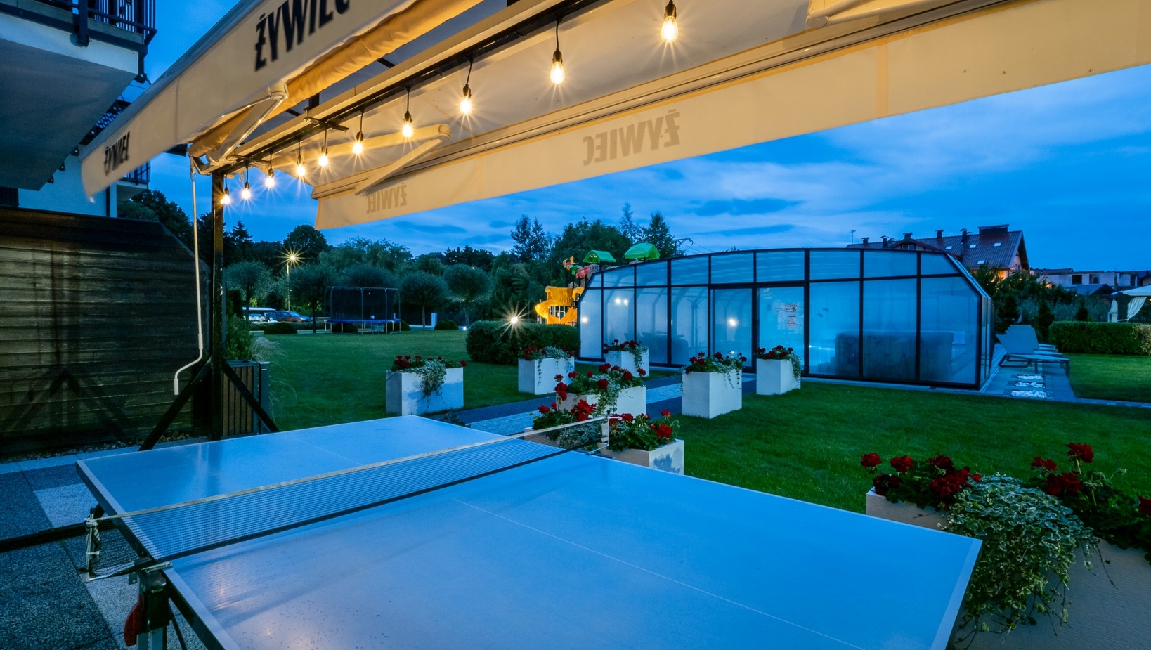 Taras hotelowy o zmierzchu z lampkami, stołem do ping-ponga, przeszklonym basenem, trawnikiem i placem zabaw