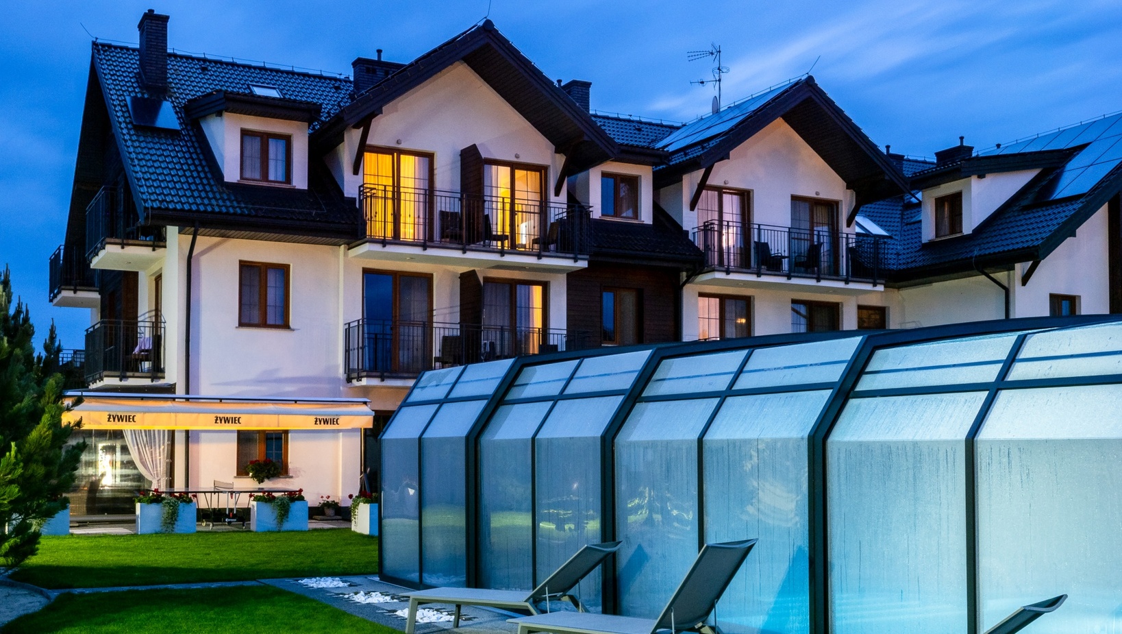 Alpine-style three-story hotel with warm-lit balconies, covered glass pool enclosure, lawn and poolside loungers at dusk.