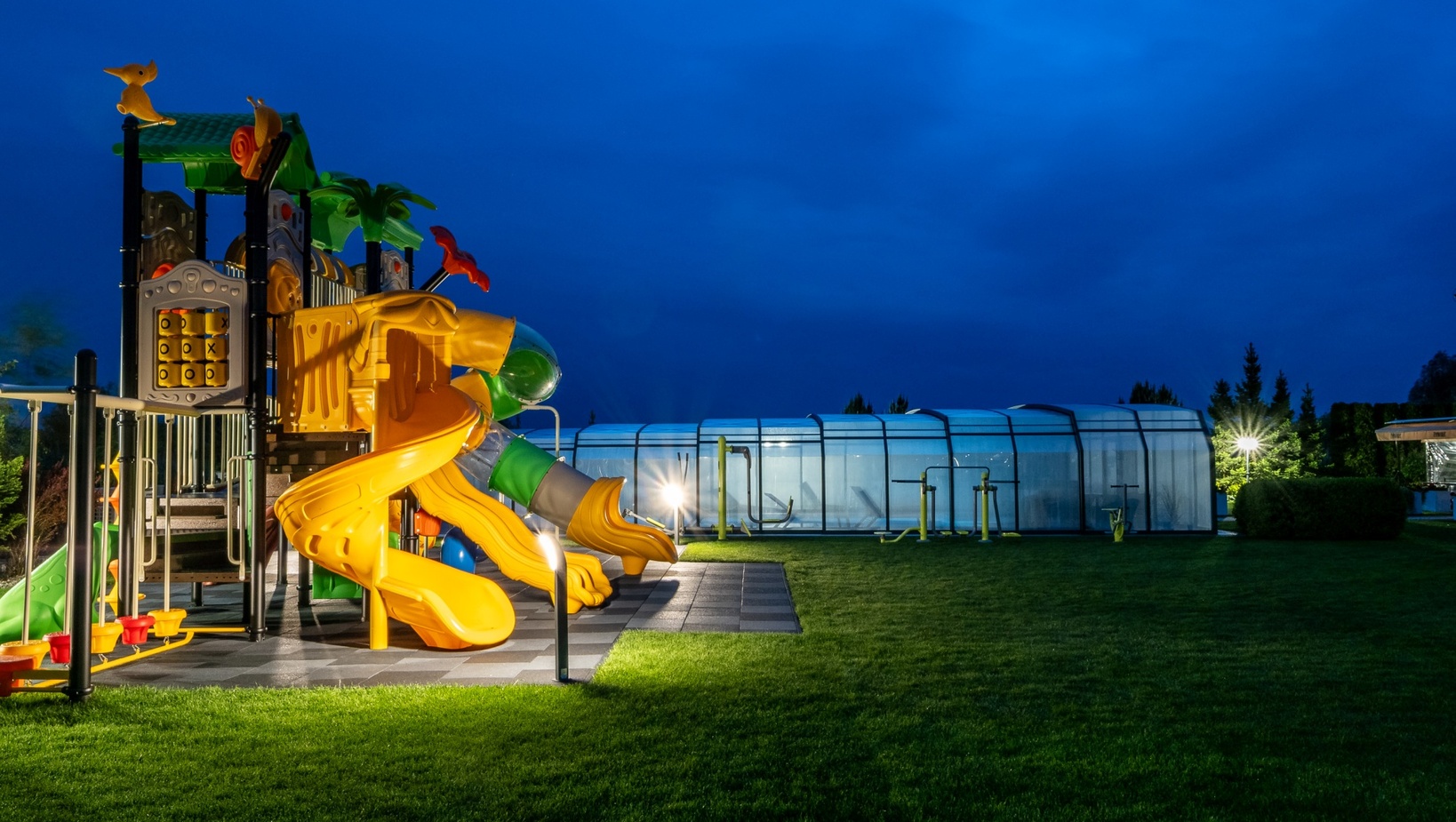 Illuminated children's playground with yellow slides on a manicured lawn beside a covered hotel pool at dusk.