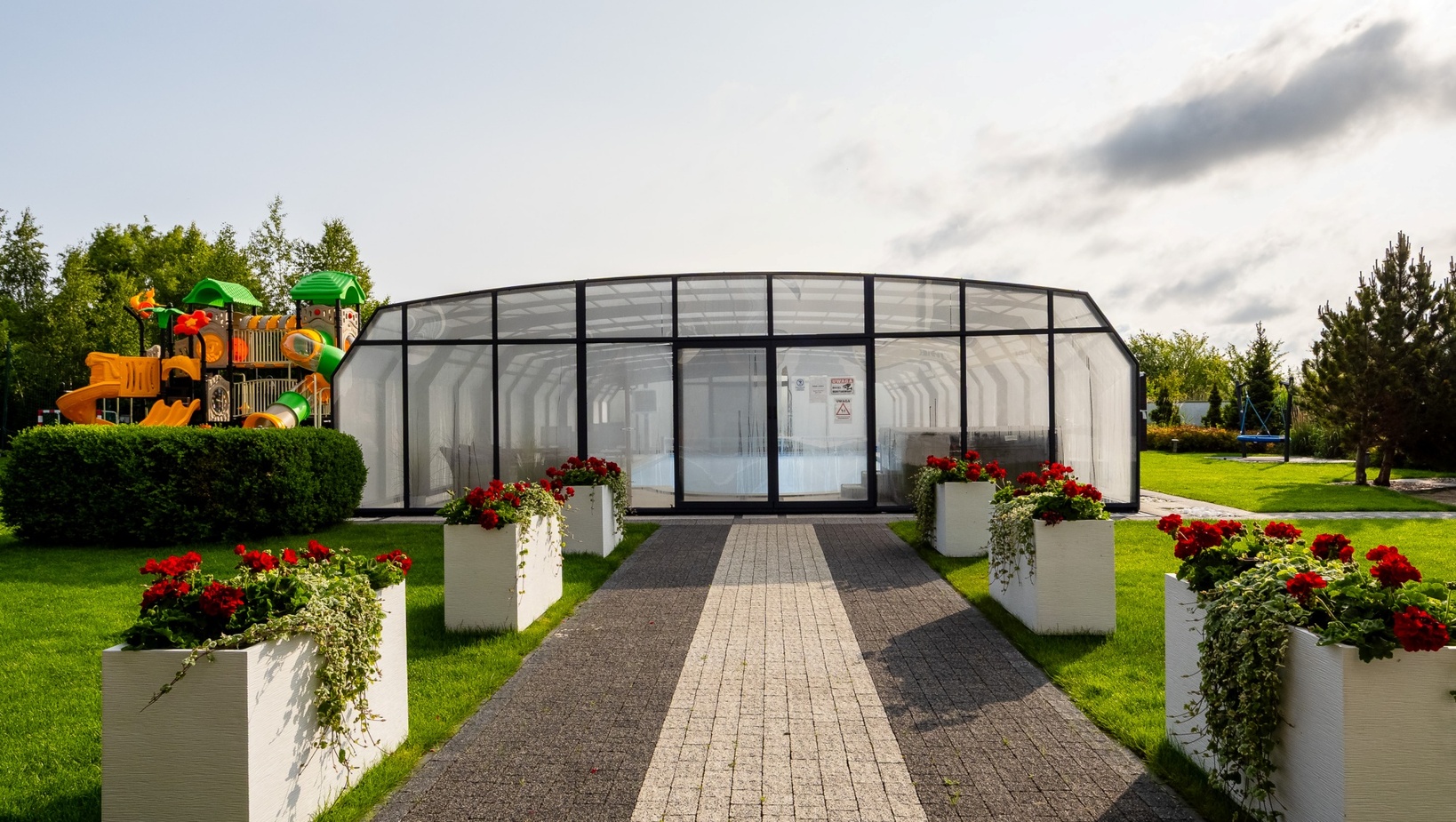 Zadaszony szklany basen z brukowanym dojściem między białymi donicami z czerwonymi kwiatami, zadbany trawnik i plac zabaw