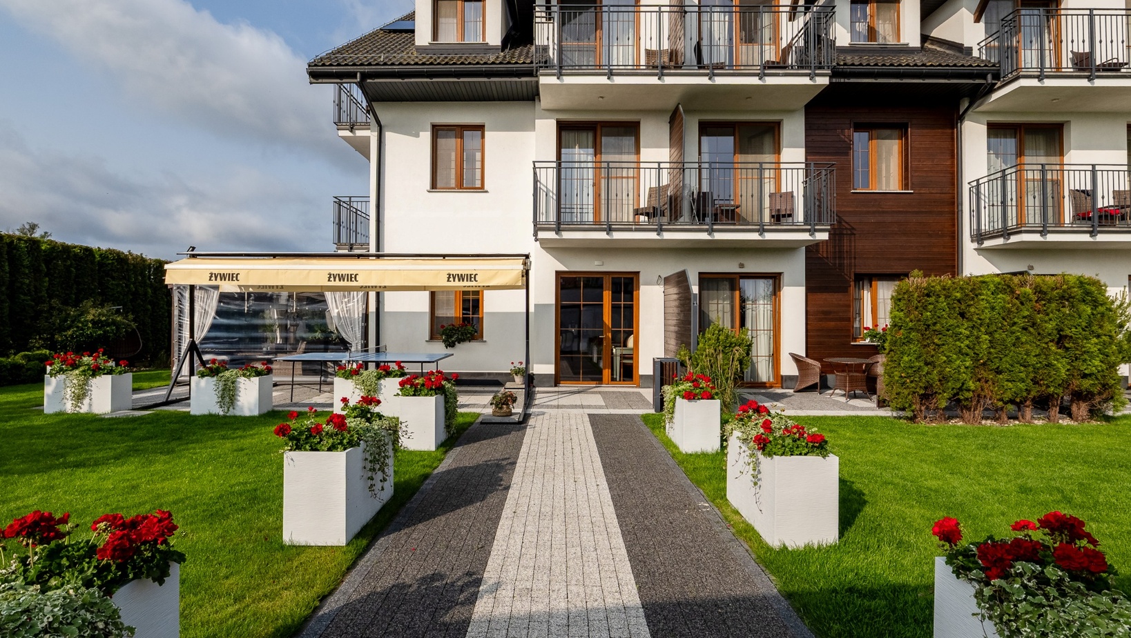 Pensjonat z balkonami, tarasem na parterze z zadaszonym miejscem do jedzenia, brukowaną ścieżką i białymi donicami z czerwonymi pelargoniami na trawniku