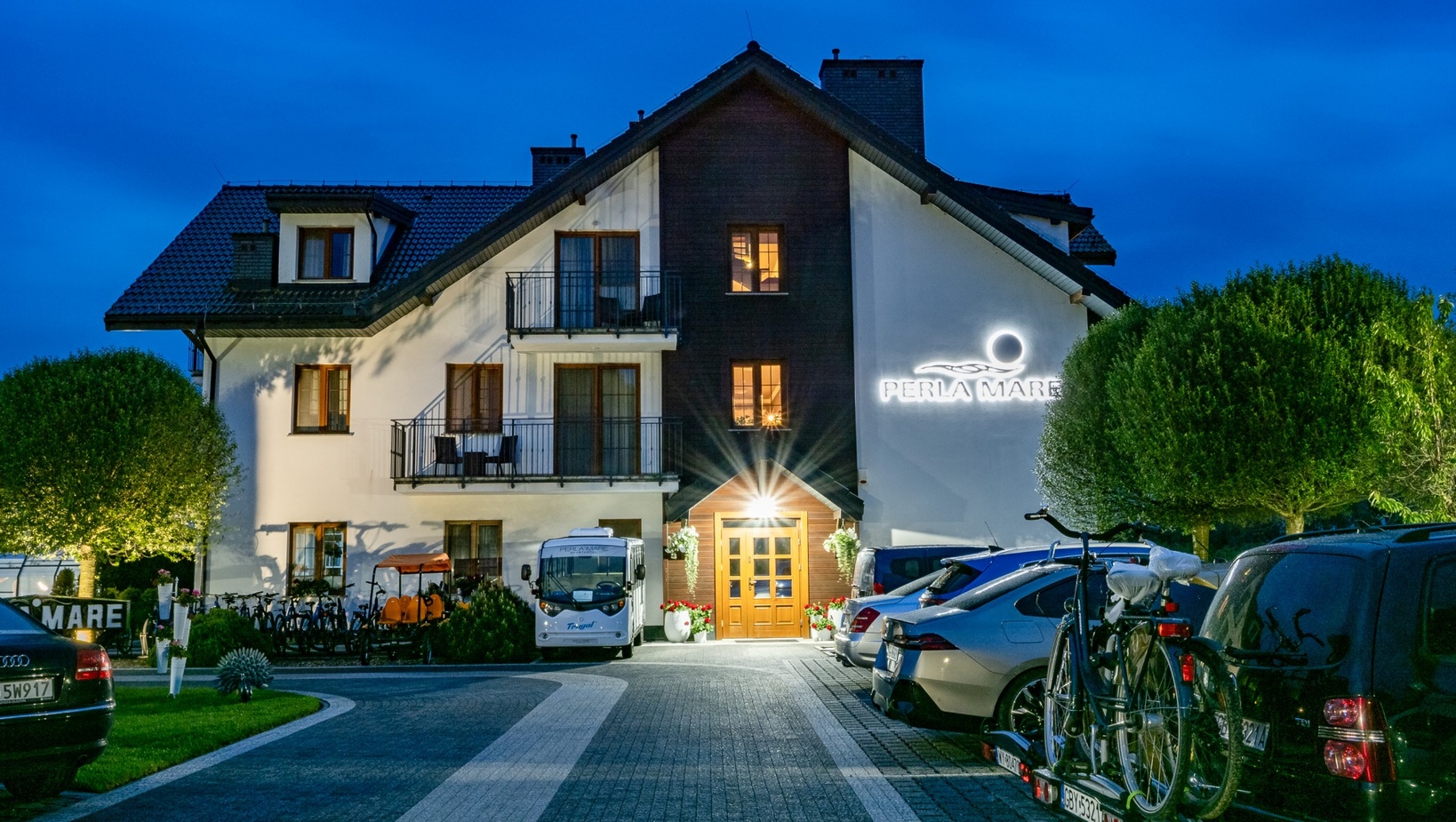 Trzypiętrowy nadmorski hotel o zmroku z podświetlonym drewnianym wejściem, balkonami, napisem „Perla Mare”, zaparkowanymi samochodami i rowerami