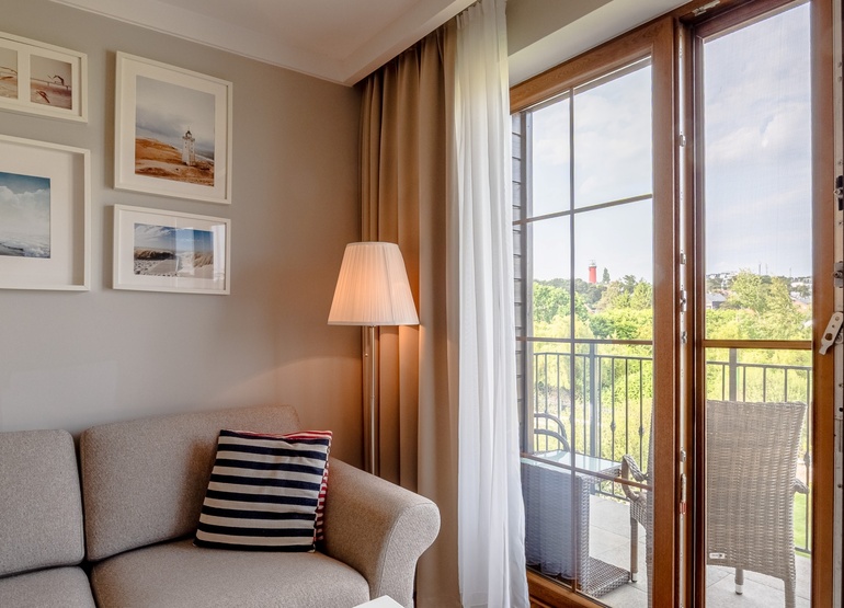 Przytulny kąt w apartamencie z beżową sofą, pasiastymi poduszkami, lampą stojącą i morskimi obrazami; prywatny balkon z rattanowymi fotelami i widokiem na latarnię