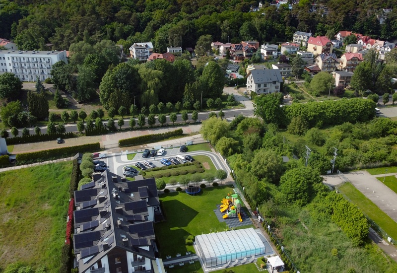 Aerial view of a guesthouse complex with solar-roofed rooms, parked cars, covered pool, children's playground and wooded hillside backdrop