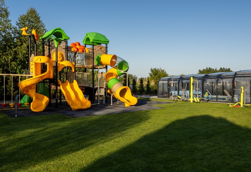 Family playground with yellow spiral and tube slides, shaded canopies on lawn beside covered pool and outdoor gym