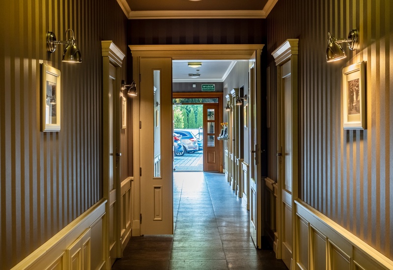 Warm-lit hotel corridor with striped wallpaper, brass wall sconces, framed art, paneled doors and view to entrance and parked cars.