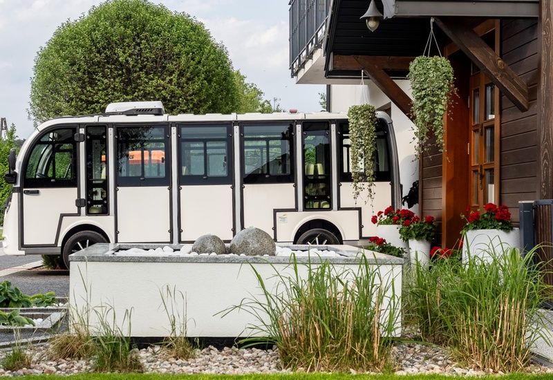 White hotel shuttle parked by a wooden entrance with hanging plants, red geraniums and decorative stone fountain
