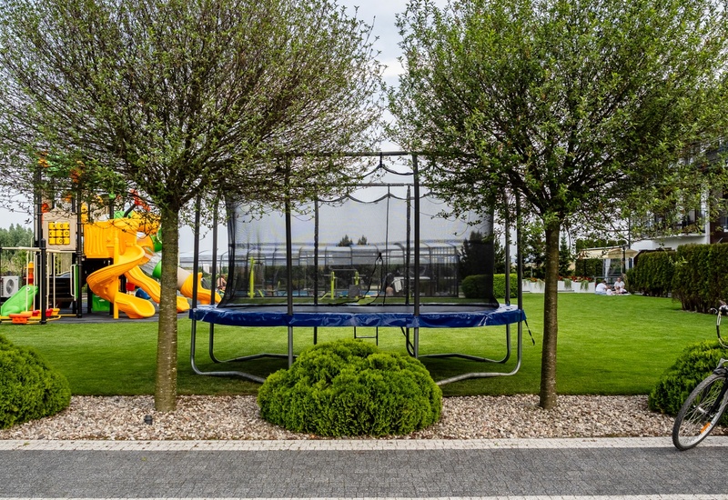 Green hotel lawn with enclosed trampoline, colorful children's playground, bicycles parked by the path and guests picnicking near the terrace.