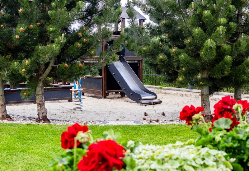 Hotel children's playground with a black slide and sandy play area framed by pine trees, green lawn and red geraniums.