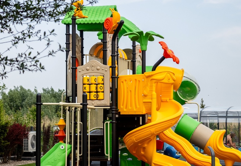 Colorful outdoor children's playground with green canopy, yellow spiral and tube slides, tic-tac-toe panel and soft artificial grass.