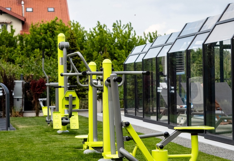 Outdoor fitness stations on a manicured lawn beside a glass-enclosed pool with sun loungers and garden views.