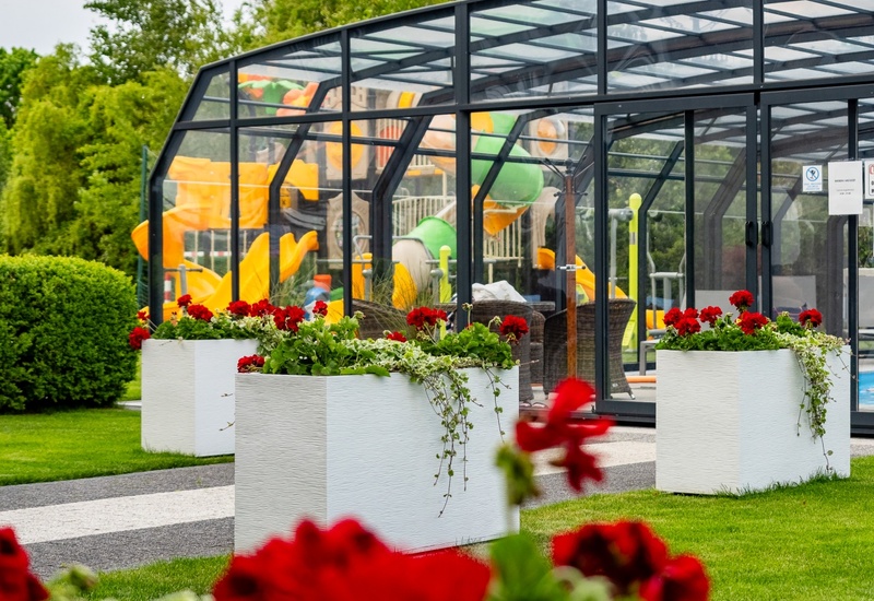 Glass-enclosed pool pavilion with colorful water slides, manicured lawn and white planters of red geraniums — family-friendly resort feature