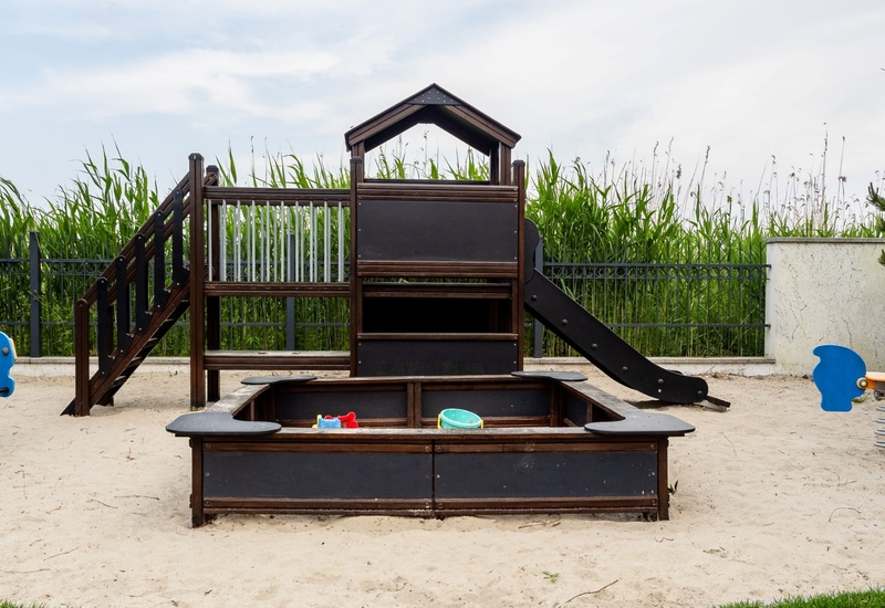 Sandy kids' play area with wooden playhouse and slide, central sandbox with toys and two blue spring riders.