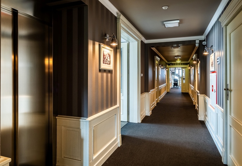 Elegant hotel corridor with carpet, white paneled wainscoting, striped wallpaper, wall sconces, framed art, elevator and stroller at far end