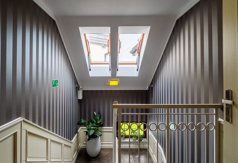 Bright hotel stair landing with twin skylights, striped walls, white paneled wainscoting, decorative railing and potted plants