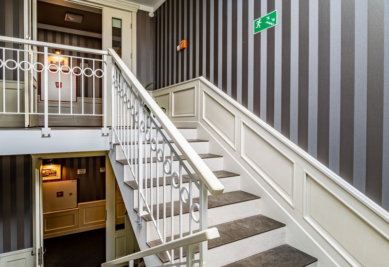 Hotel stairwell with white railing and circular motifs, striped wallpaper, paneled wainscot and illuminated corridor.