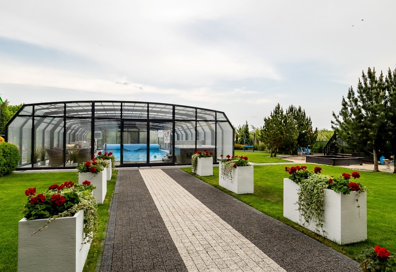 Paved walkway with white planters and red flowers leading to a glass-enclosed pool; green lawns, pines and children's playground.