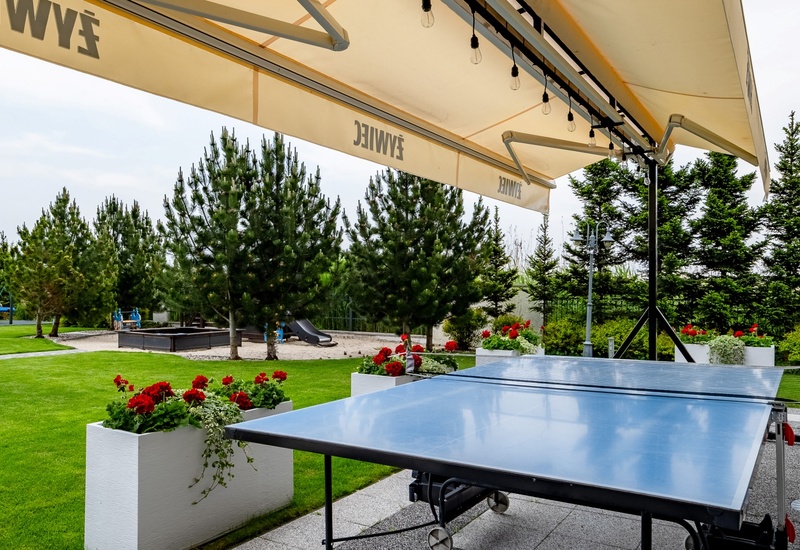 Outdoor ping-pong table under a large terrace awning, manicured lawn, red flower boxes and a children's playground among pine trees.