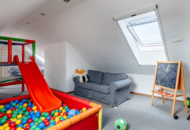 Hotel children's playroom with colorful ball pit and red slide, cozy gray sofa under skylight, chalkboard easel and toys.